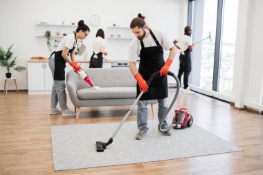 Professional bearded cleaner from cleaning service vacuums carpet using wireless vacuum cleaner against background of multinational colleagues cleaning bright, modern kitchen. clipart