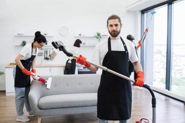 Kablosuz elektrik süpürgesini elinde tutarken gülümseyen sakallı temizlikçinin portresi, kameraya bakıyor, arka planda çok kültürlü meslektaşların parlak, modern mutfağı temizliyor..