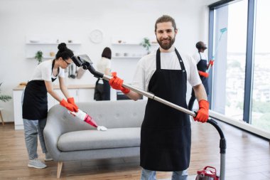 Kablosuz elektrik süpürgesini elinde tutarken gülümseyen sakallı temizlikçinin portresi, kameraya bakıyor, arka planda çok kültürlü meslektaşların parlak, modern mutfağı temizliyor..