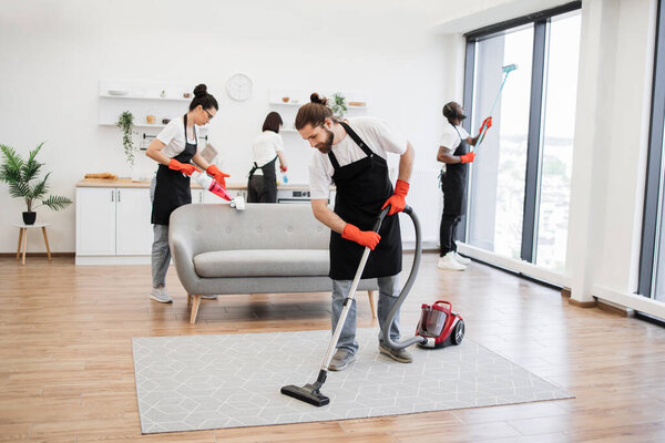 Team of professional people from cleaning service cleans sofa, carpet, cuisine table and panoramic windows of spacious apartment. Multinational group of cleaners cleans bright studio kitchen.