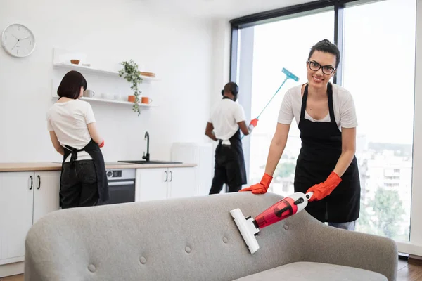 Happy smiling employees of cleaning company cleaning in bright, spacious home kitchen. Multiethnic young female cleaner vacuuming sofa with portable cordless vacuum cleaner.