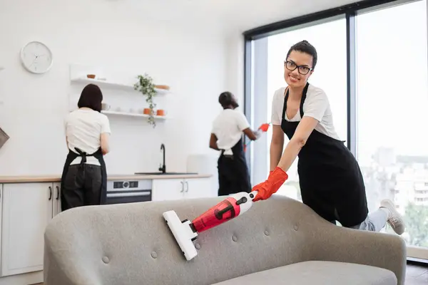 Happy smiling employees of cleaning company cleaning in bright, spacious home kitchen. Multiethnic young female cleaner vacuuming sofa with portable cordless vacuum cleaner.