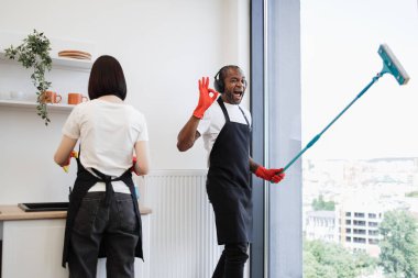 Parlak modern tavan arası mutfağını panoramik pencereli temizleyen çok ırklı şirket çalışanları. Genç Afrikalı pencereleri paspasla yıkıyor, kulaklıkla müzik dinliyor ve işaret diliyle dans ediyor..