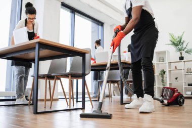 Elektrikli süpürge tutan Afrikalı adamın elleri. Genç çok kültürlü temizlikçilerden oluşan bir ekip yerleri süpürür, masaları ve aletleri siler, rafları, geniş, parlak, modern ofislerin camlarını siler..