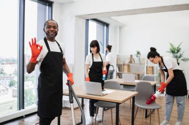 Çok kültürlü temizlikçiler masaları, aletleri, rafları silerler. Geniş, parlak, modern ofisin camlarını silerler. Afro-Amerikalı adam elektrik süpürgesiyle yerleri temizliyor..