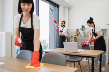 Siyah önlüklü, kırmızı lastik eldivenli, panoramik pencere camını paspasla temizleyen, modern ofiste çok ırklı iş arkadaşlarıyla kameraya bakan pozitif sakallı beyaz işçiye odaklan..