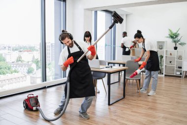 Profesyonel temizlikçi, elinde elektrikli süpürge tutarken konserde çalan gitar taklidi yapar gibi eğleniyor. Modern ofisi temizleyen siyah önlüklü, çok uluslu mutlu takım..