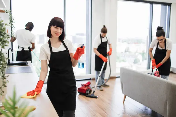 Cleaning service team at work in kitchen in private home. Caucasian janitor in apron cleaning table with detergent and gloves while her multiethnic colleague wiping floor, windows and vacuuming sofa.