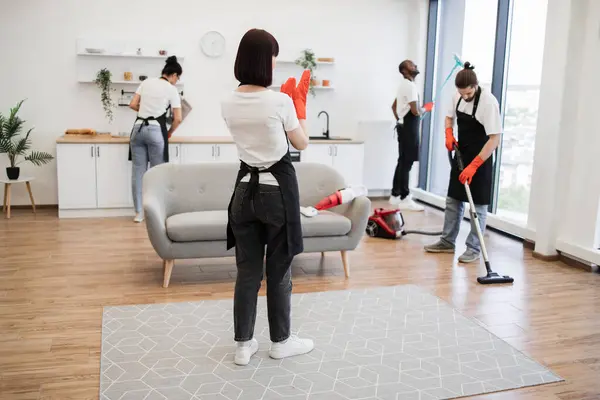 Female boss of company distributes tasks to employees and points to cleaning locations. Professional housekeeper services company team working at customer house washing dishes, kitchen cleaning.