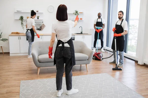 Professional housekeeper services company team working at customer house washing dishes, kitchen cleaning. Female boss of company distributes tasks to employees and points to cleaning locations.