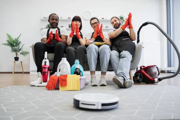 Robot vacuum cleaner cleans floor while team of professional housekeepers resting and clapping hands on couch. Multiracial cleaners in black uniform and red gloves relaxing after work on sofa.