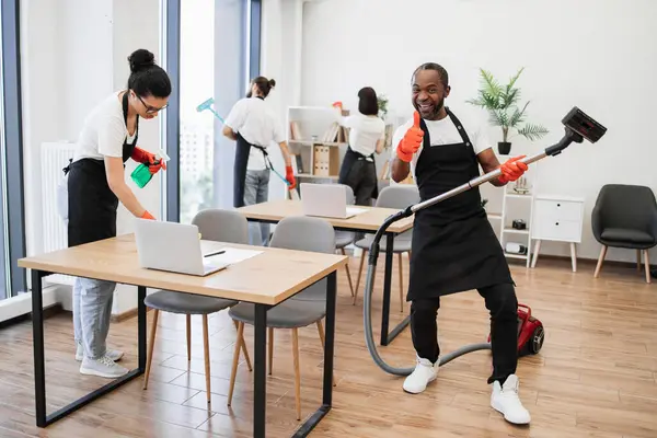 Professional cleaner African man having fun holding vacuum cleaner like guitar imitating playing at concert showing thumb up. Happy multinational team of people in black aprons cleaning modern office.
