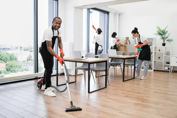 Professional service team of cleaning company. Young adult African American man wearing black apron and red rubber gloves vacuuming floor of modern light office.