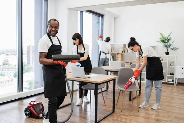 Team of multicultural cleaners wipes tables and gadgets, shelves, washes windows in spacious, bright, modern office. Side view of African American man using vacuum cleaner to clean floor from dirt.
