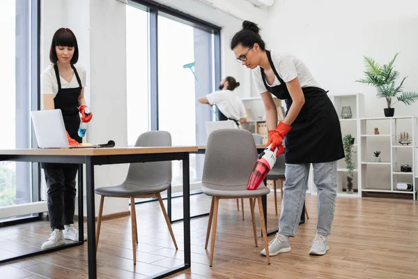 Multiethnic young female cleaner vacuuming chair with portable cordless vacuum cleaner. Happy smiling multicultural employees of company cleaning in bright, spacious modern office.