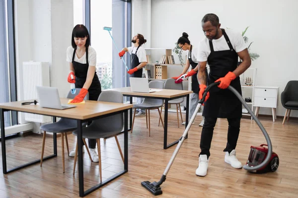 Professional team of people of cleaning service in black aprons and red gloves cleans tables, floor, and panoramic windows of spacious studio . Multinational group cleans bright office.