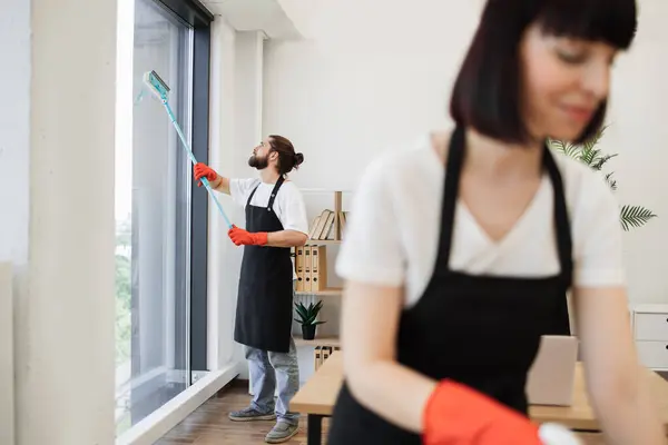 Professional team of people cleaning service in black aprons and red gloves cleans tables, floor, and panoramic windows of spacious studio . Multinational group cleans bright office.