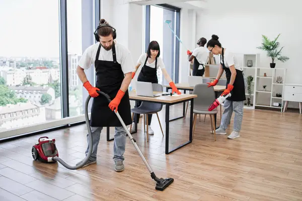 Team of young multicultural cleaners vacuums floor, wipes tables with gadgets, shelves, washes windows in spacious, bright, modern office. Professional cleaning company.