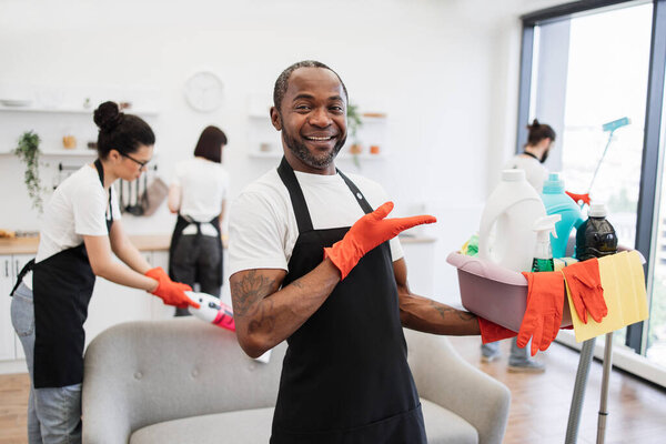 Portrait of young African American man professional cleaning worker pointing on bucket for washing with detergents on bright kitchen studio background, copy space.