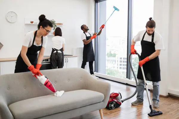 Professional team of people of cleaning service cleans sofa, carpet, cuisine table and panoramic windows of spacious apartment. Multinational group of cleaners cleans bright studio kitchen.