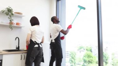 Afro-Amerikan işçilerin panoramik pencere camlarını paspasla temizlemelerine, kameraya bakmalarına, modern dairede çok ırklı iş arkadaşlarının yanında başparmağını göstermelerine odaklan..