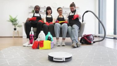 Profesyonel temizlikçiler dinlenirken ve koltukta el çırparken robot süpürgeyle yerleri temizlemeye odaklan. Siyah üniformalı çok ırklı temizlikçiler ve işten sonra koltukta dinlenen kırmızı eldivenler.