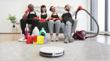 Profesyonel temizlikçiler kanepede oturup gülümseyip dinlenirken modern robot süpürgesi yerleri temizliyor. Siyah üniformalı çok ırklı temizlikçiler ve işten sonra koltukta dinlenen kırmızı eldivenler.