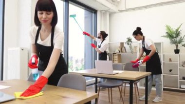 Siyah önlüklü ve kırmızı eldivenli profesyonel temizlik ekibi geniş stüdyonun masalarını, zeminlerini ve panoramik pencerelerini temizler. Çokuluslu grup parlak ofisi temizliyor.