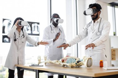 Working atmosphere in modern lab. Concentrated multiethnic professional biologists, doctors, scientists wearing VR glasses working with human skeleton on the table.