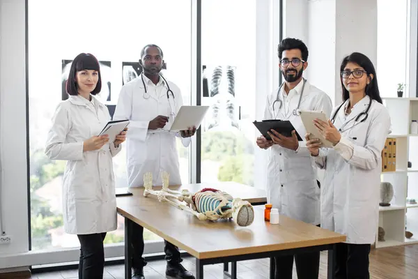 Young multiethnic medical students on anatomy lesson in modern classroom looking at camera. Middle aged male doctor professor teaching anatomy using human skeleton model with with body organs.