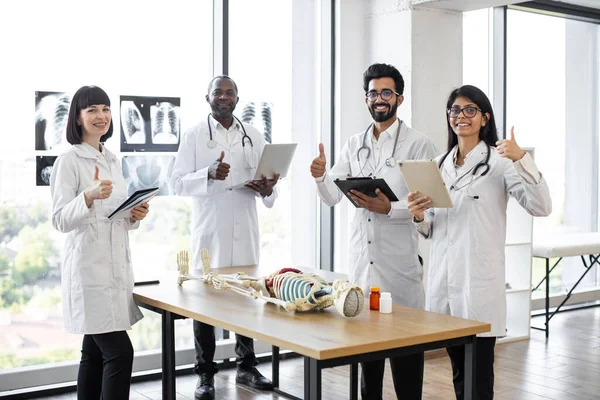 Young multiethnic medical students on anatomy lesson in modern classroom showing thumbs up. Middle aged male doctor professor teaching anatomy using human skeleton model with with body organs.