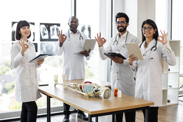 Young multiethnic medical students on anatomy lesson in modern classroom showing sign ok. Middle aged male doctor professor teaching anatomy using human skeleton model with with body organs.