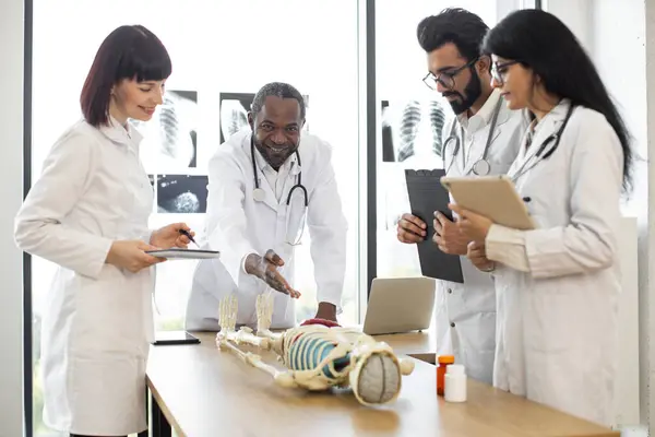 African man medical teacher teaching anatomy to university students or young doctors. Arabian man, Caucasian and Indian women standing near table, working with human skeleton model with body organs.