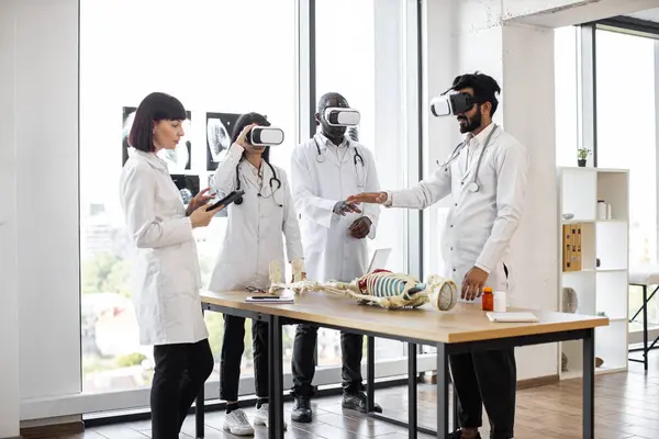 Science, technology, virtual reality and people concept. Group of multiethnic medical students or workers using virtual reality goggles to exam human skeleton lying on the table.