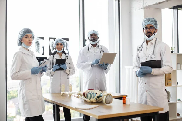 Multiethnic medical students in protective masks, gloves and hats on anatomy lesson in classroom looking at camera. Middle aged doctor teaching anatomy using human skeleton model with with body organs