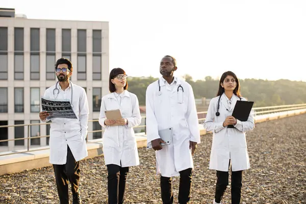 Group of multiracial relaxed doctors walking together outdoors on rooftop terrace of modern medical clinic. Successful meeting of medical workers team of international hospital.