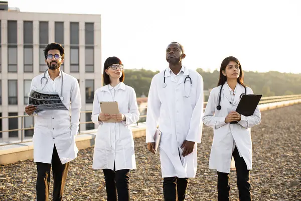 Group of multiracial relaxed doctors walking together outdoors on rooftop terrace of modern medical clinic. Successful meeting of medical workers team of international hospital.