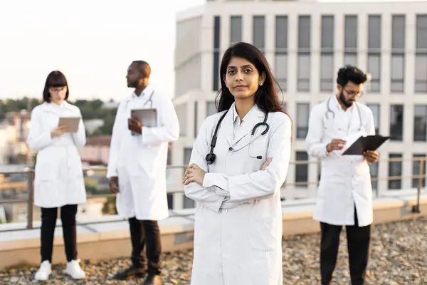 Portrait of confident young Indian doctor in white lab coat posing on camera with crossed hands and smiling. International colleagues of mixed nationality standing behind and operating modern gadgets.