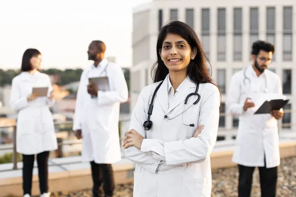 Portrait of confident young Indian doctor in white lab coat posing on camera with crossed hands and smiling. International colleagues of mixed nationality standing behind and operating modern gadgets.