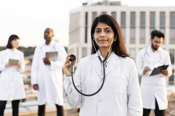 Portrait of confident young Indian doctor in white lab coat posing on camera holding a stethoscope outdoors. International colleagues of mixed nationality standing behind and operating modern gadgets.