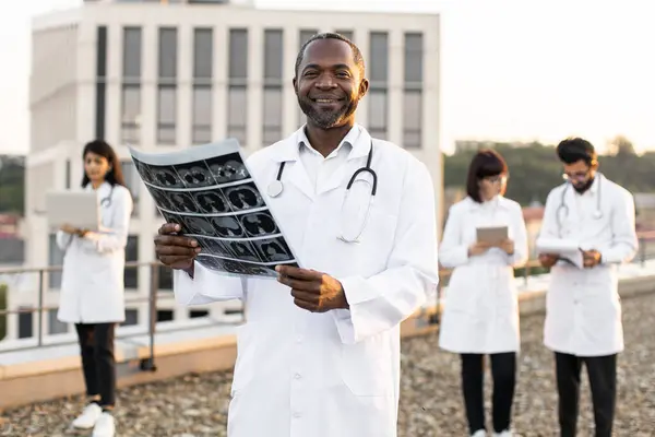Portrait of African doctor holding x-ray scan of patient during break outdoors hospital looking at camera. Multiethnic employees of clinic working on devices with results of examination of patients.