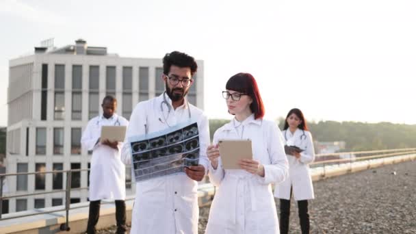 国际医疗工作者团体在城市的屋顶 身穿白色实验室外套 留着胡子的医生向年轻的白人护士展示核磁共振X光 同时两名同事在室外休息时放松一下 — 图库视频影像