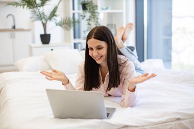 Gülümseyen bir kadının, rahat bir yatakta yatarken bilgisayarında arkadaşlarıyla görüntülü konuşma görüntüsü. Çekici kadın, evde kalırken modern teknolojileri kullanarak uzaktan iletişimden keyif alıyor..