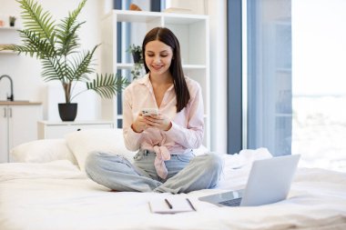 Rahat bir kadın rahat bir yatakta oturuyor ve hafta sonları akıllı telefon kullanarak iş arkadaşlarıyla sohbet ediyor. Evden uzakta çalışan çekici genç bir kadın modern kablosuz cep telefonuyla daktilo çekiyor..