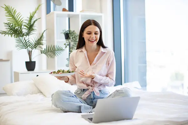 Charming woman in casual attire enjoying breakfast of fresh vegetables while sitting on cozy bed, staying at home. Happy brunette female talking by video call using laptop and eating healthy salad.