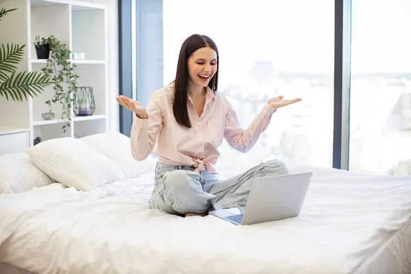 Charming woman in casual attire enjoying online conversation with friends while sitting on cozy bed. Happy brunette female gesturing while talking by video call using modern laptop, staying at home.