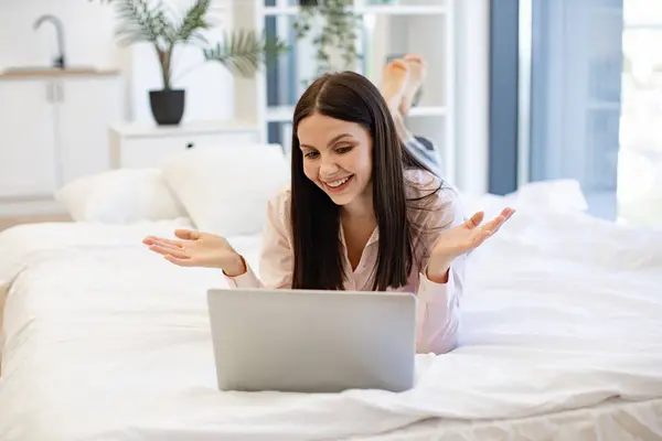 Charming woman in casual attire enjoying online conversation with friends while lying on cozy bed. Happy brunette female gesturing while talking by video call using modern laptop, staying at home.