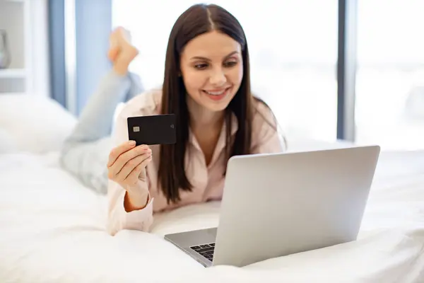 Charming woman in casual attire enjoying online shopping while lying on cozy bed. Attractive brunette female typing on modern laptop entering credit card information for purchase, staying at home.