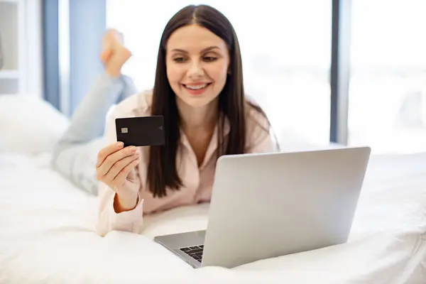 Charming woman in casual attire enjoying online shopping while lying on cozy bed. Attractive brunette female typing on modern laptop entering credit card information for purchase, staying at home.