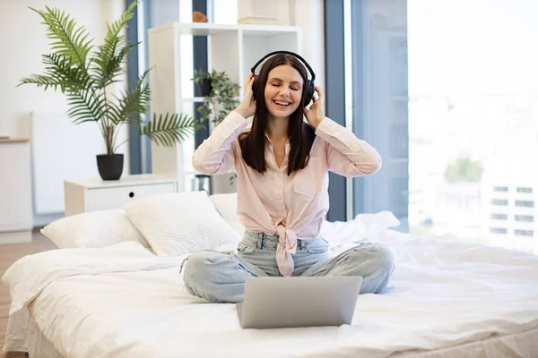 Charming woman in casual attire dancing of favorite music during break of remote work while sitting on cozy bed. Smiling brunette female in wireless headphones enjoying favorite song, staying at home.
